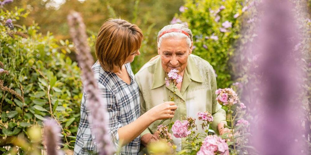 Eine Frau hält einer Seniorin in einem Garten eine Blume Richtung Nase. Die Seniorin riecht an der Blume.