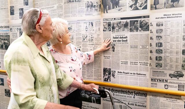 Eine Seniorin und eine Frau gehen einen Flur entlang, welcher mit Zeitungen von früher tapeziert ist. Die Seniorin geht an einem Gehwagen und beide Frauen schauen sich die Zeitungen an.  