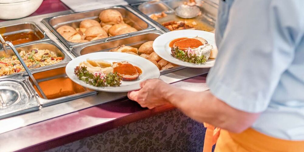 Eine Servicekraft steht am Essensbüfett und hält zwei gefüllte Teller in der Hand. Hinter dem Tresen steht jemand und befüllt Teller.